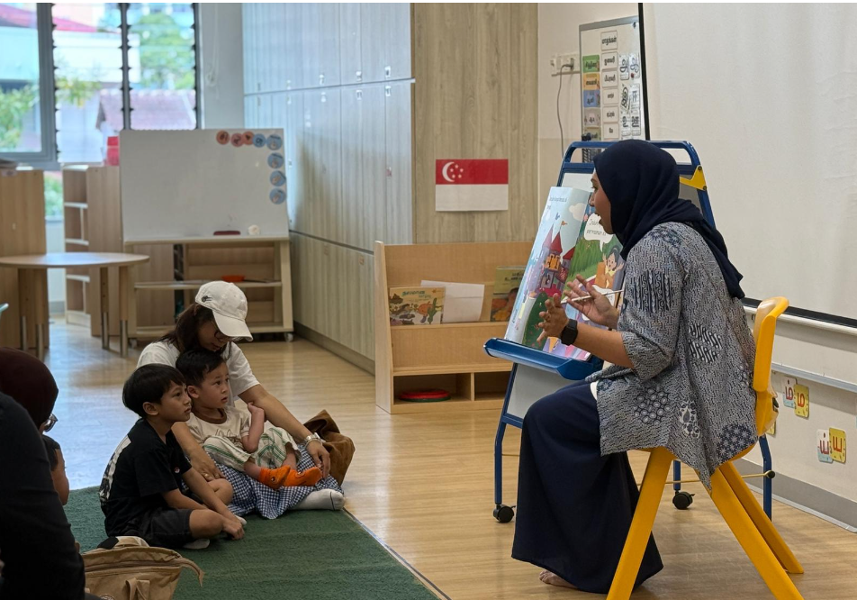 Teacher in headscarf reads from an easel book to children seated on a classroom rug.
